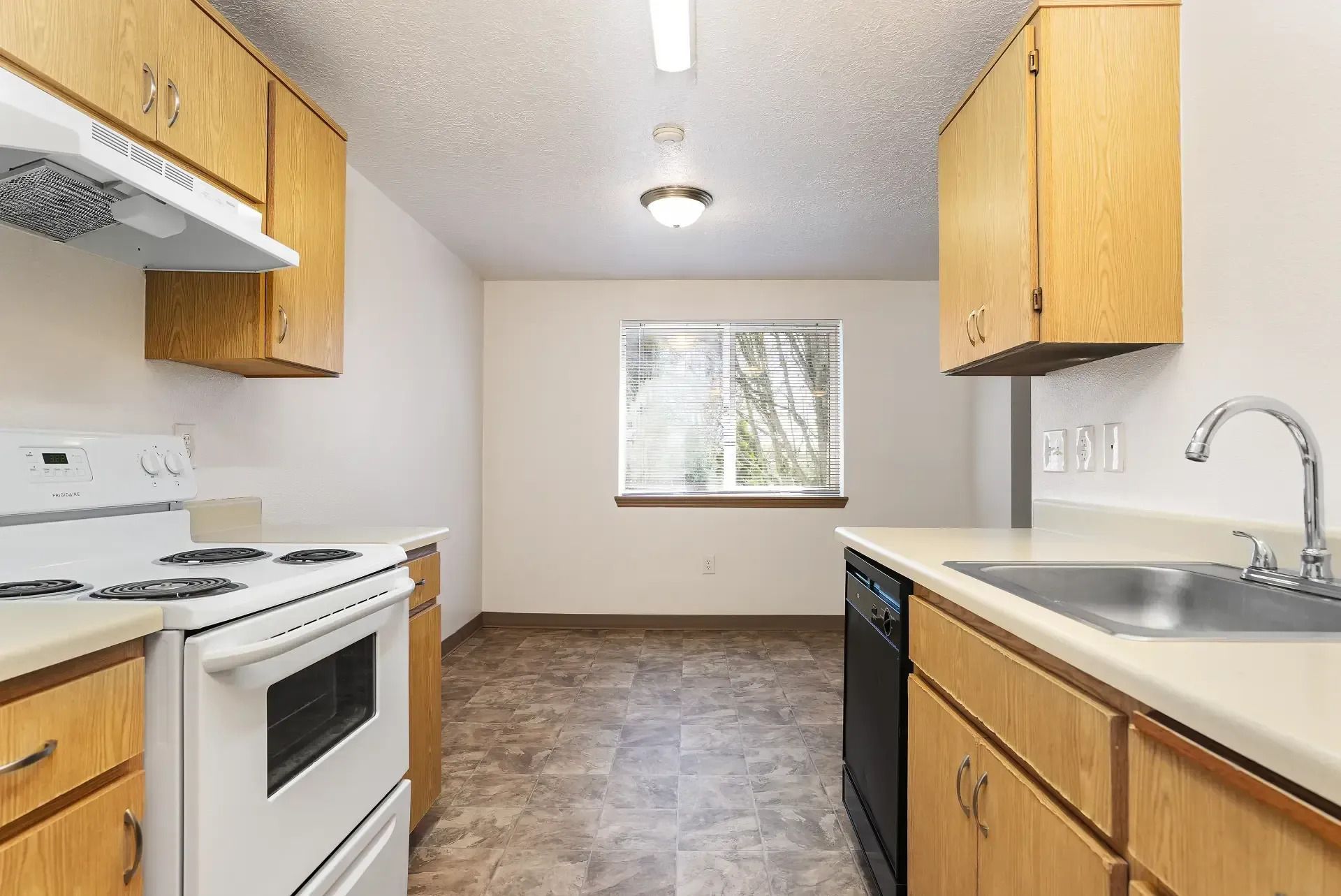 Small white kitchen with wood cabinets, white stove and sink, and a window over dark flooring