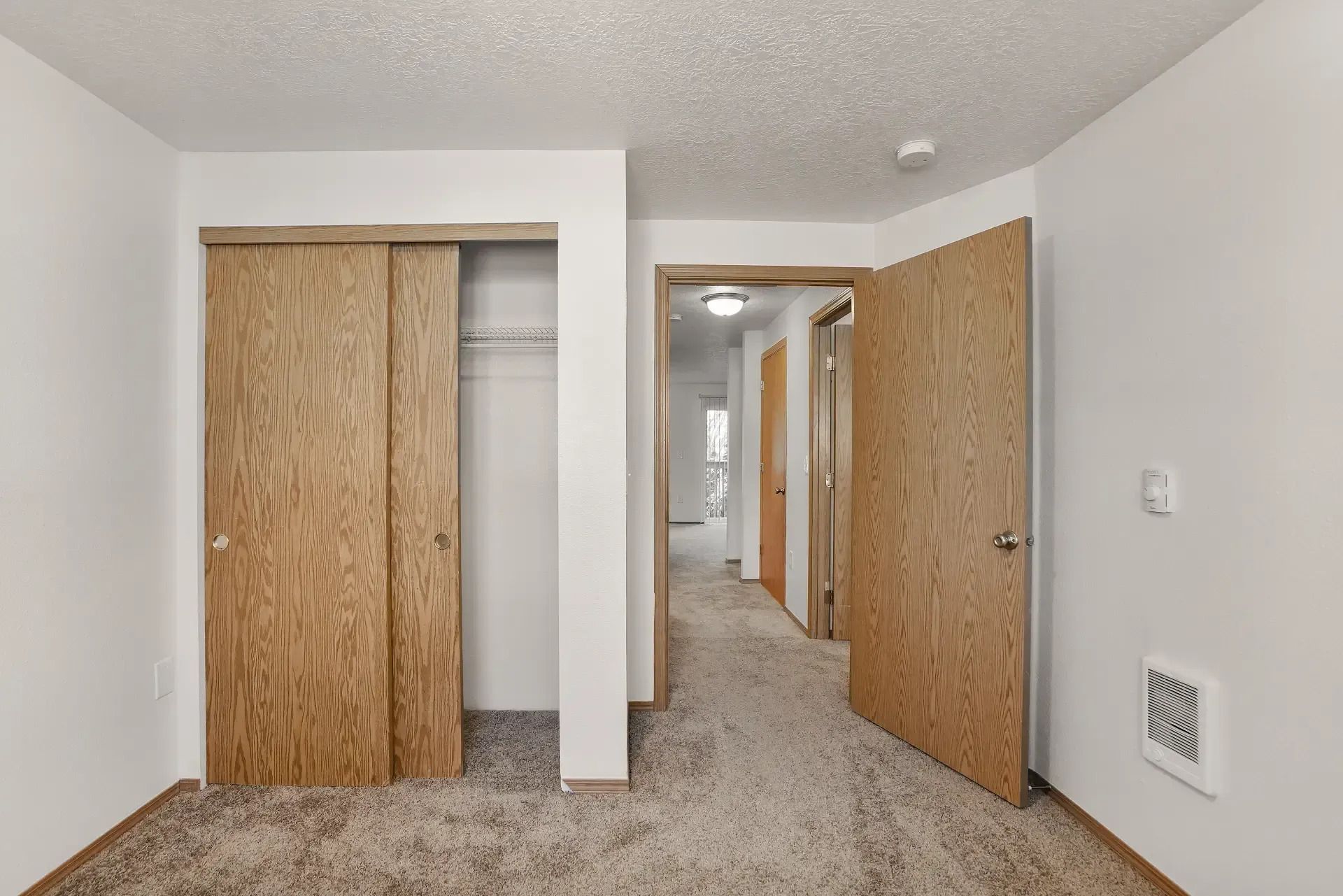 Empty carpeted bedroom with open wooden closet doors and a hallway beyond
