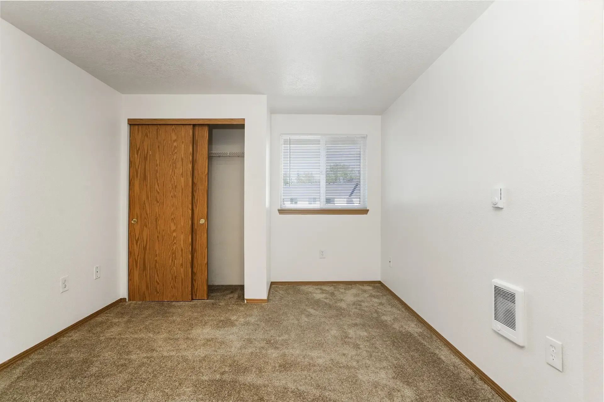 Empty bedroom with beige carpet, white walls, closet, and window with blinds.