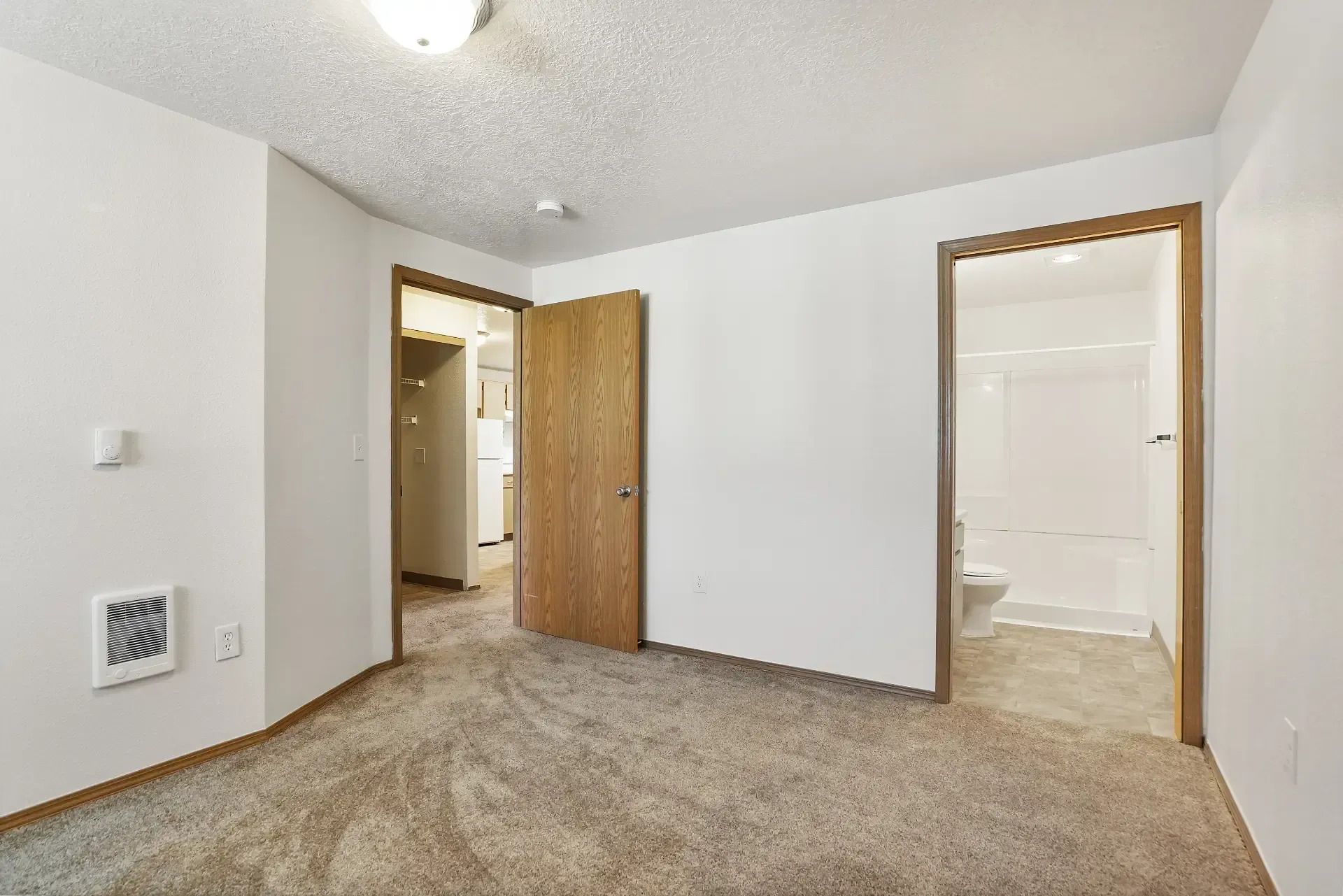 Empty carpeted bedroom with white walls, open wooden doors, and an adjoining bathroom visible.