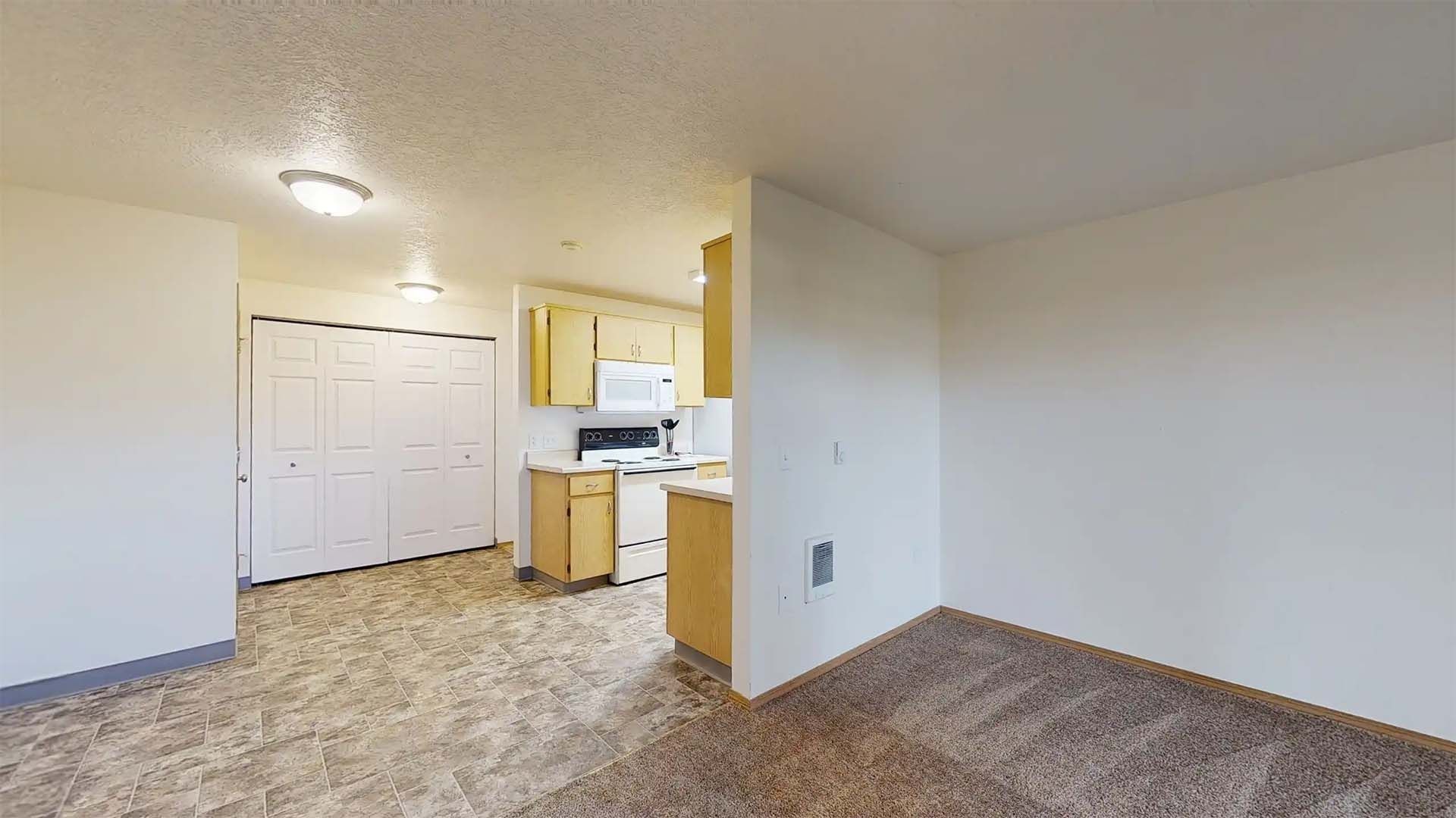 Empty apartment living area with kitchen nook, white walls, and beige carpet