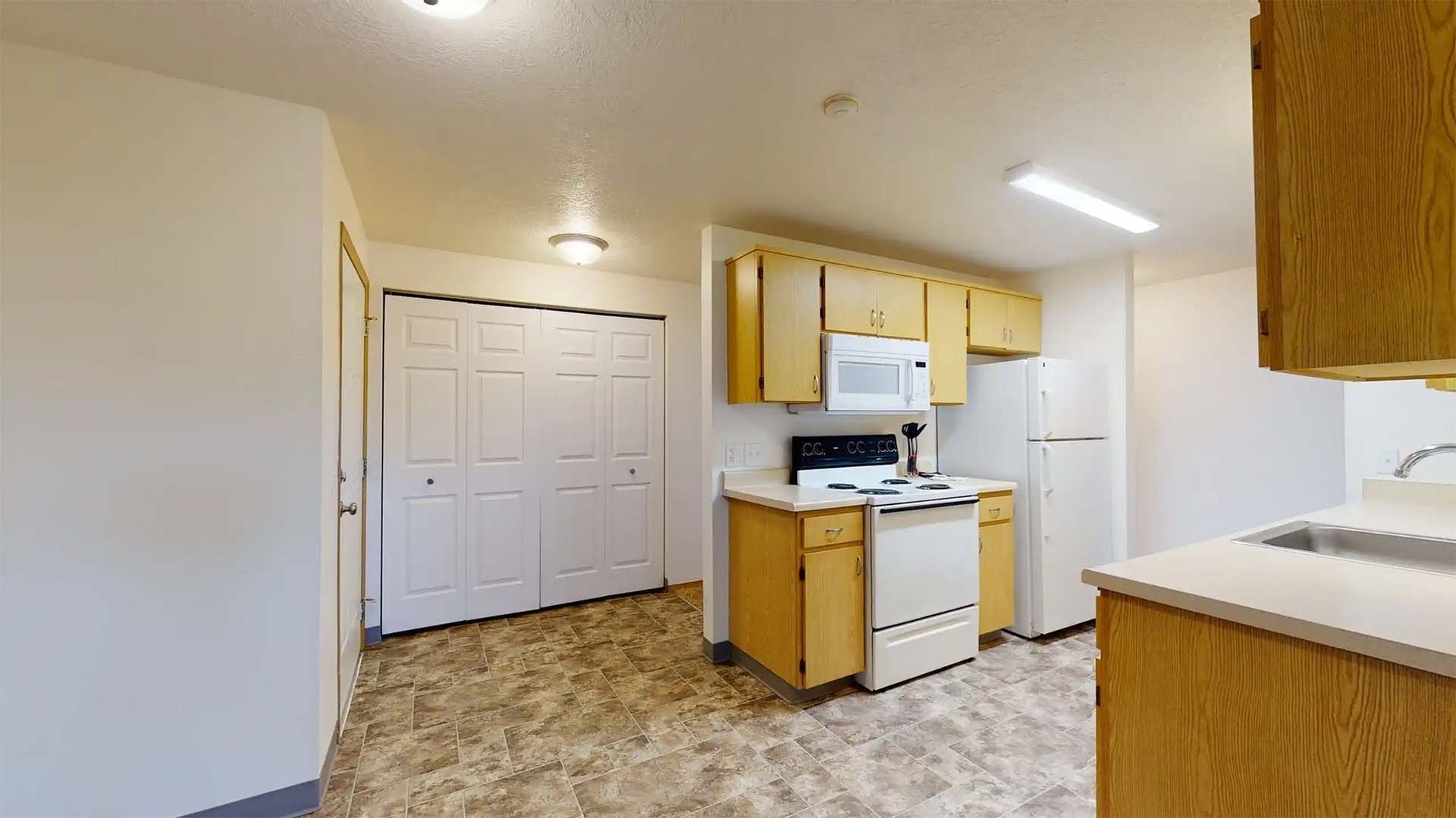 Small kitchen with white appliances, wood cabinets, and beige tile flooring near a hallway.