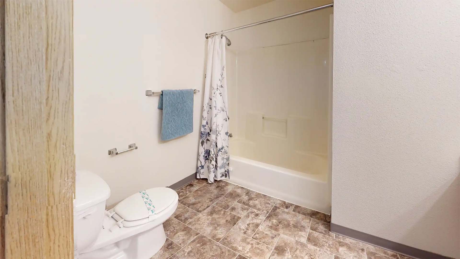 Small bathroom with toilet, bathtub, blue towel, and floral shower curtain.