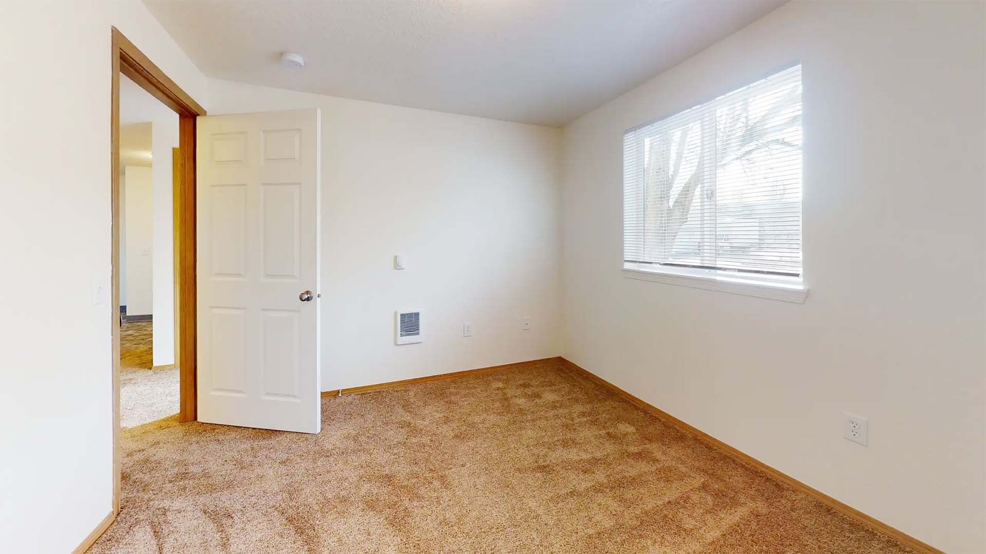 Empty beige bedroom with tan carpet, white walls, open door, and sunlit window with blinds
