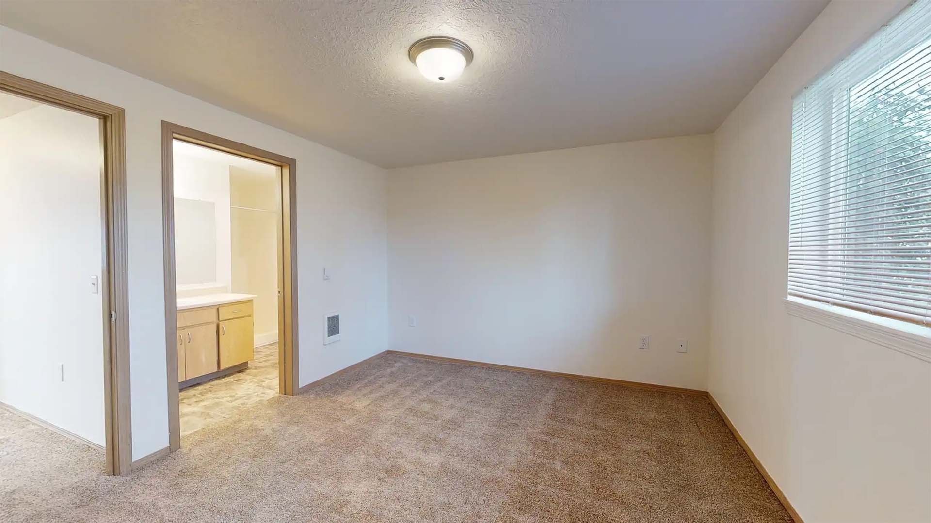 Empty beige bedroom with carpet, window blinds, open doorway to bathroom, and ceiling light