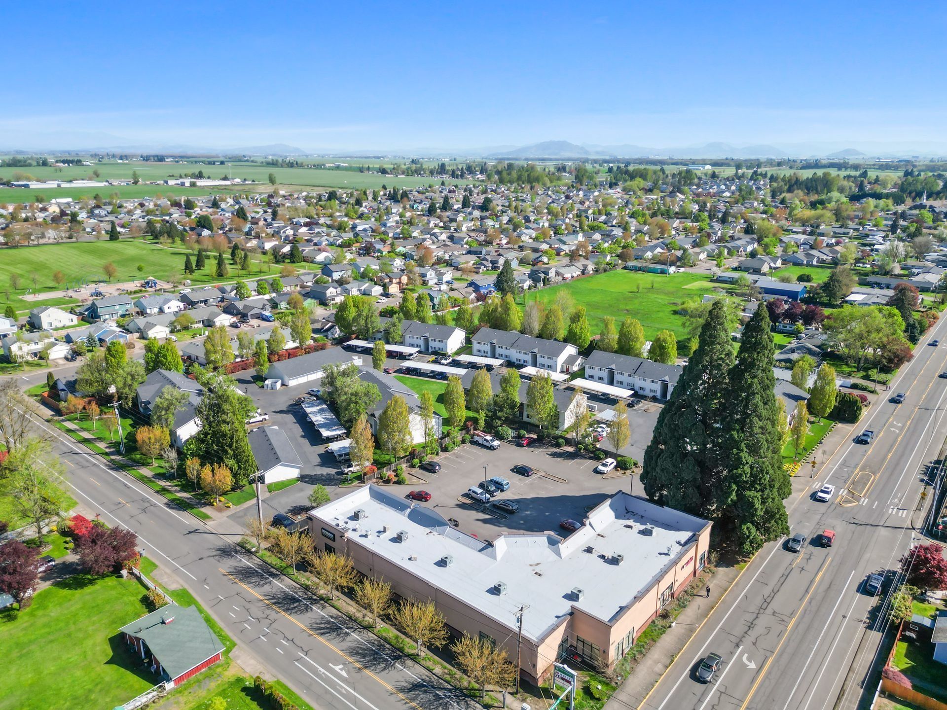 Aerial view of a suburban neighborhood with houses, trees, roads, and a large white-roofed building on a sunny day