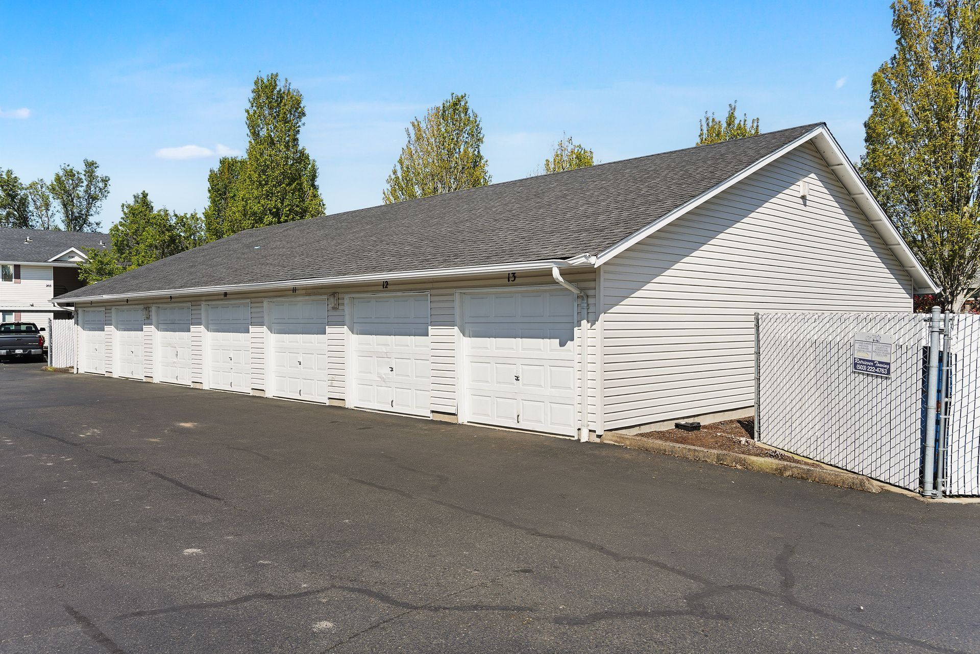 White garage row with pitched roof beside a paved driveway and chain-link fence under a clear blue sky
