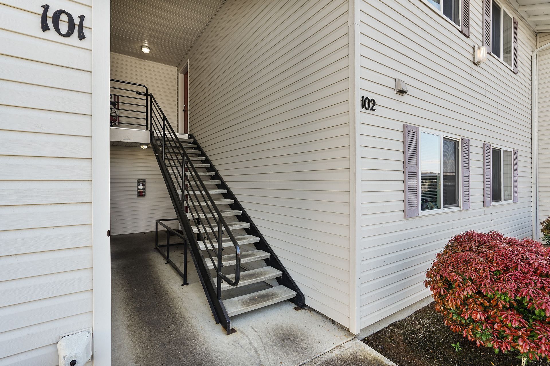 Exterior stairway beside a white apartment building with black railings and a red shrub