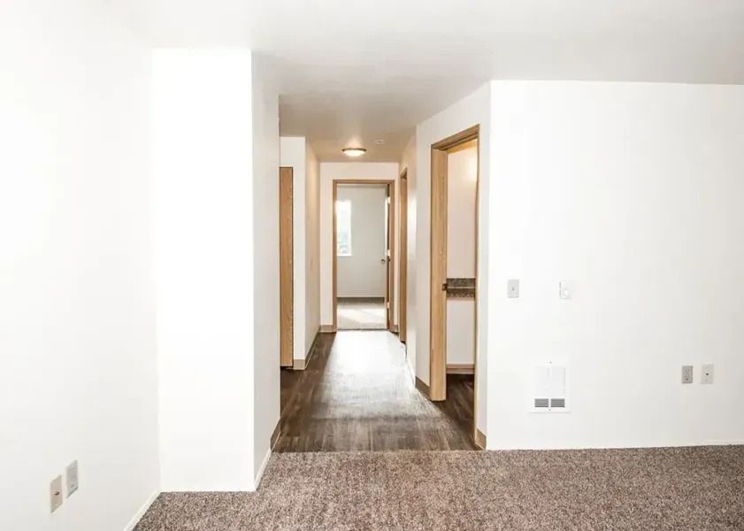 Bright hallway with white walls, wood flooring, and doorways leading into a carpeted room.