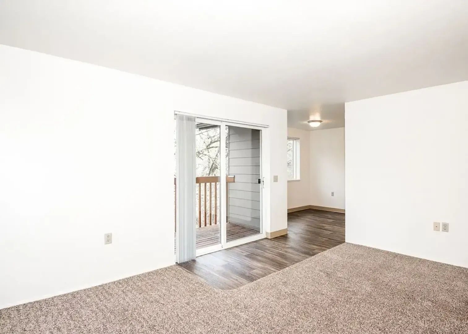 Empty carpeted living room with white walls, sliding glass doors, and a hallway in the background