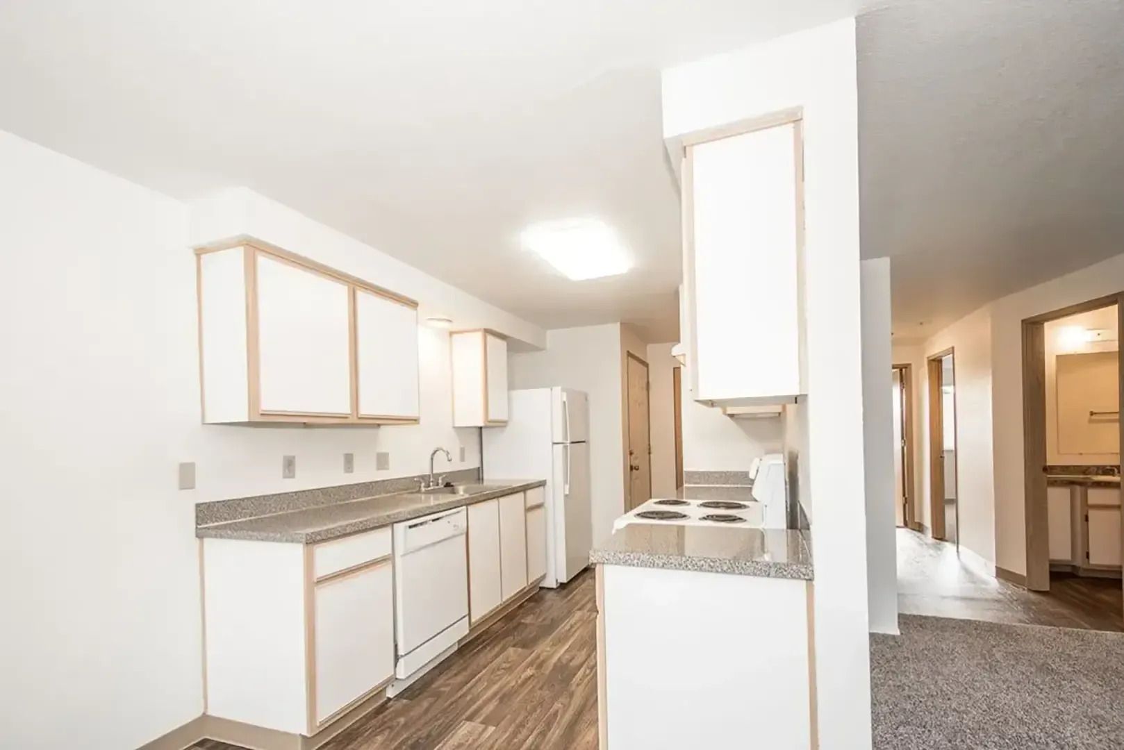 Bright white kitchen with wood floors, gray countertops, and a hallway leading to carpeted rooms