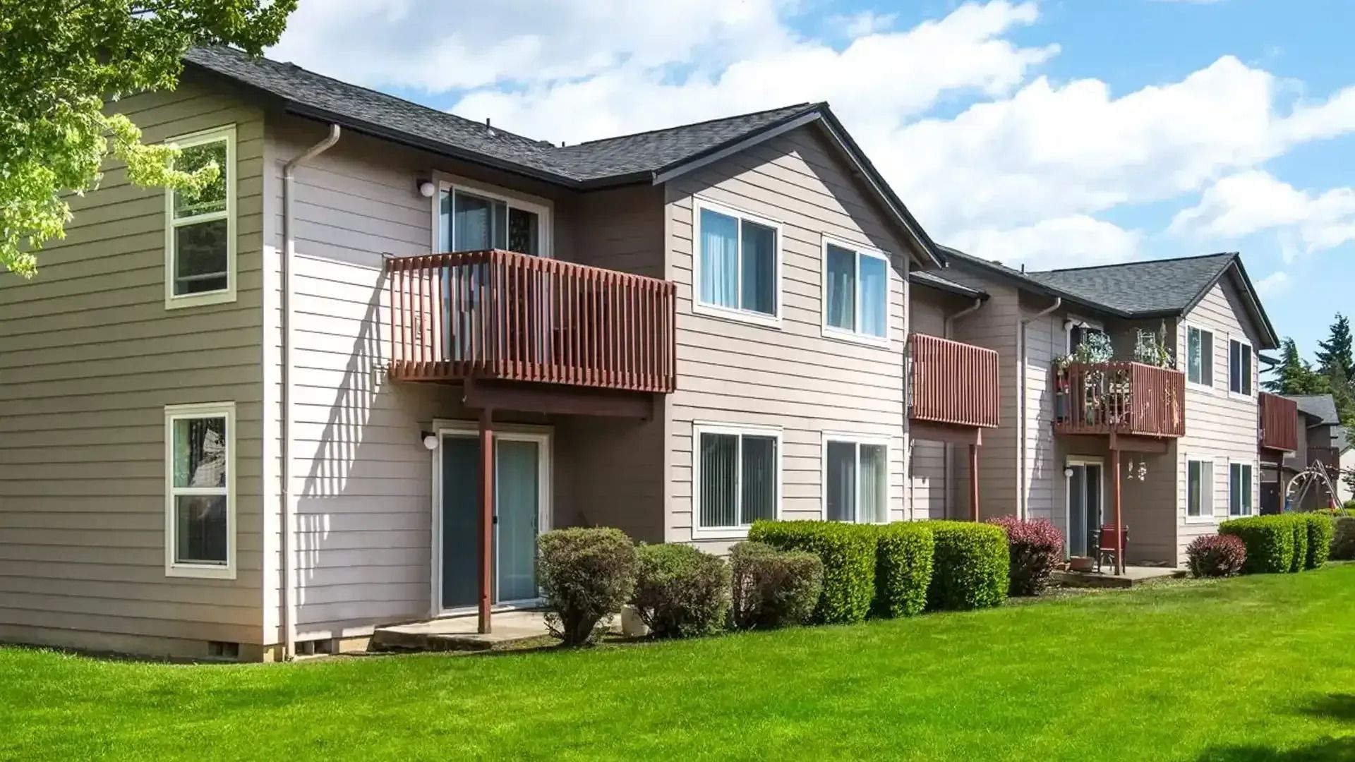 Two-story apartment building with beige siding, balconies, and green lawn under a partly cloudy sky