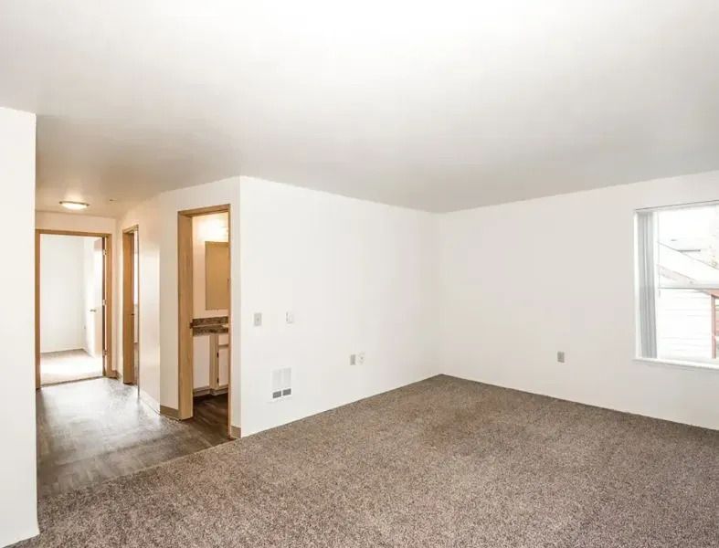 Empty beige living room with carpet, white walls, and hall and kitchen doorways visible