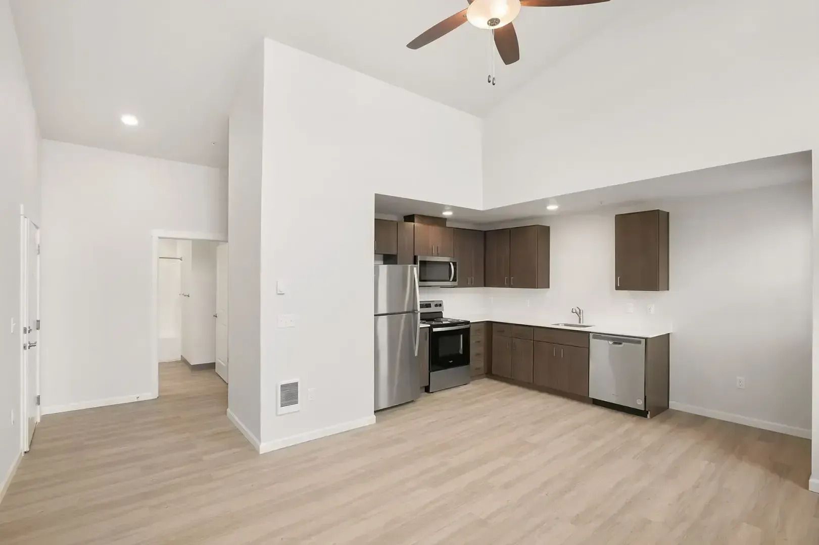 Bright empty apartment with open kitchen, light wood floors, white walls, and ceiling fan.