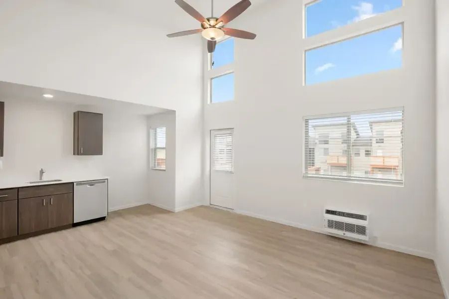 Bright empty living room with high windows, light wood floors, and a ceiling fan above a small kitchenette.