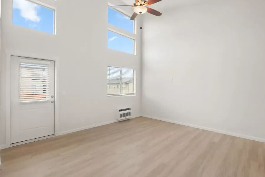 Bright empty room with high windows, white walls, light wood floor, and a ceiling fan.