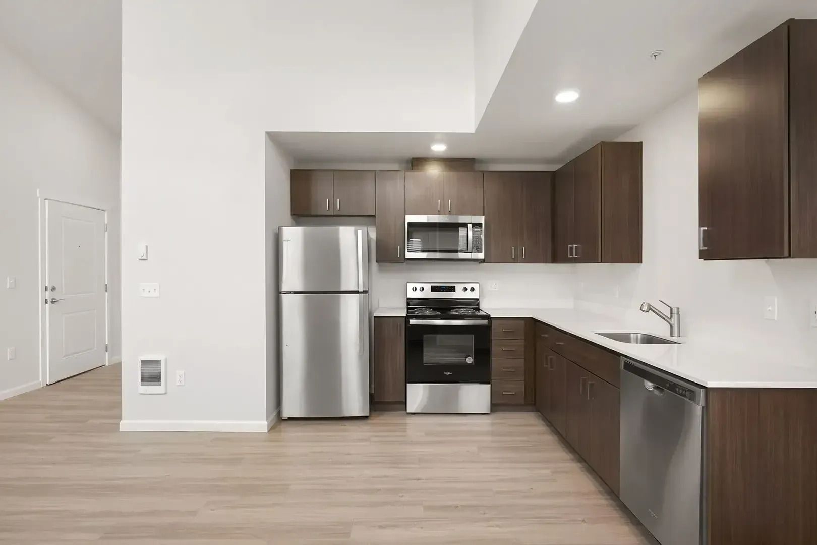 Modern kitchen with stainless steel appliances, dark wood cabinets, white walls, and light wood flooring