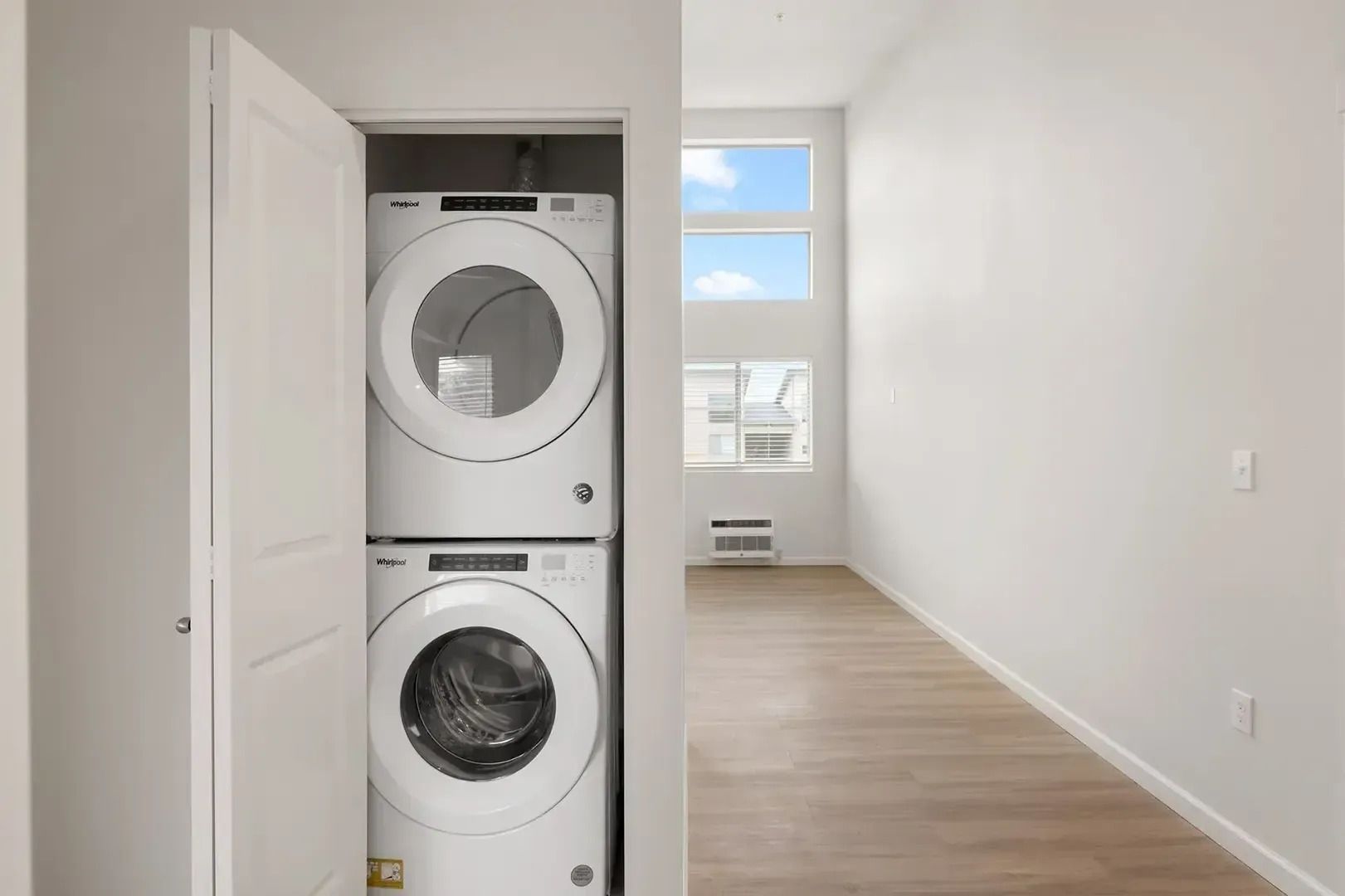Stacked washer and dryer in a narrow closet beside an empty bright room with wood flooring.