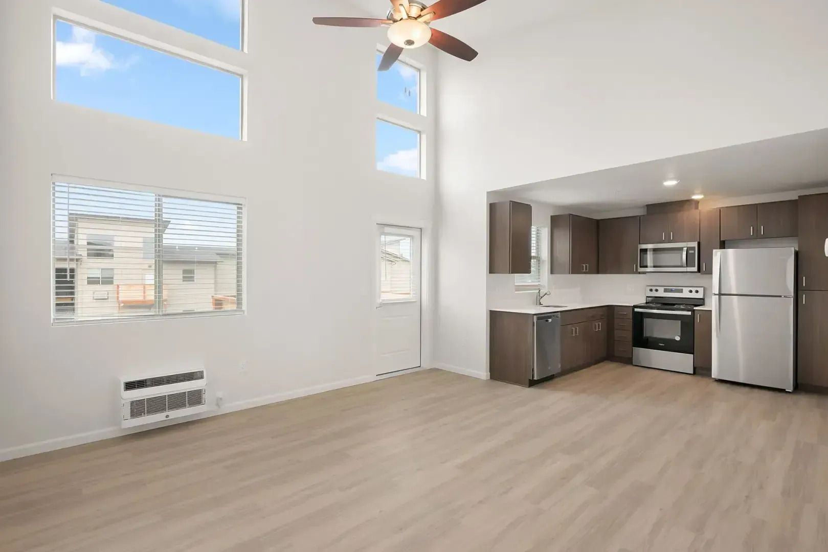 Bright empty studio apartment with high ceilings, large windows, and a compact kitchen with stainless steel appliances.