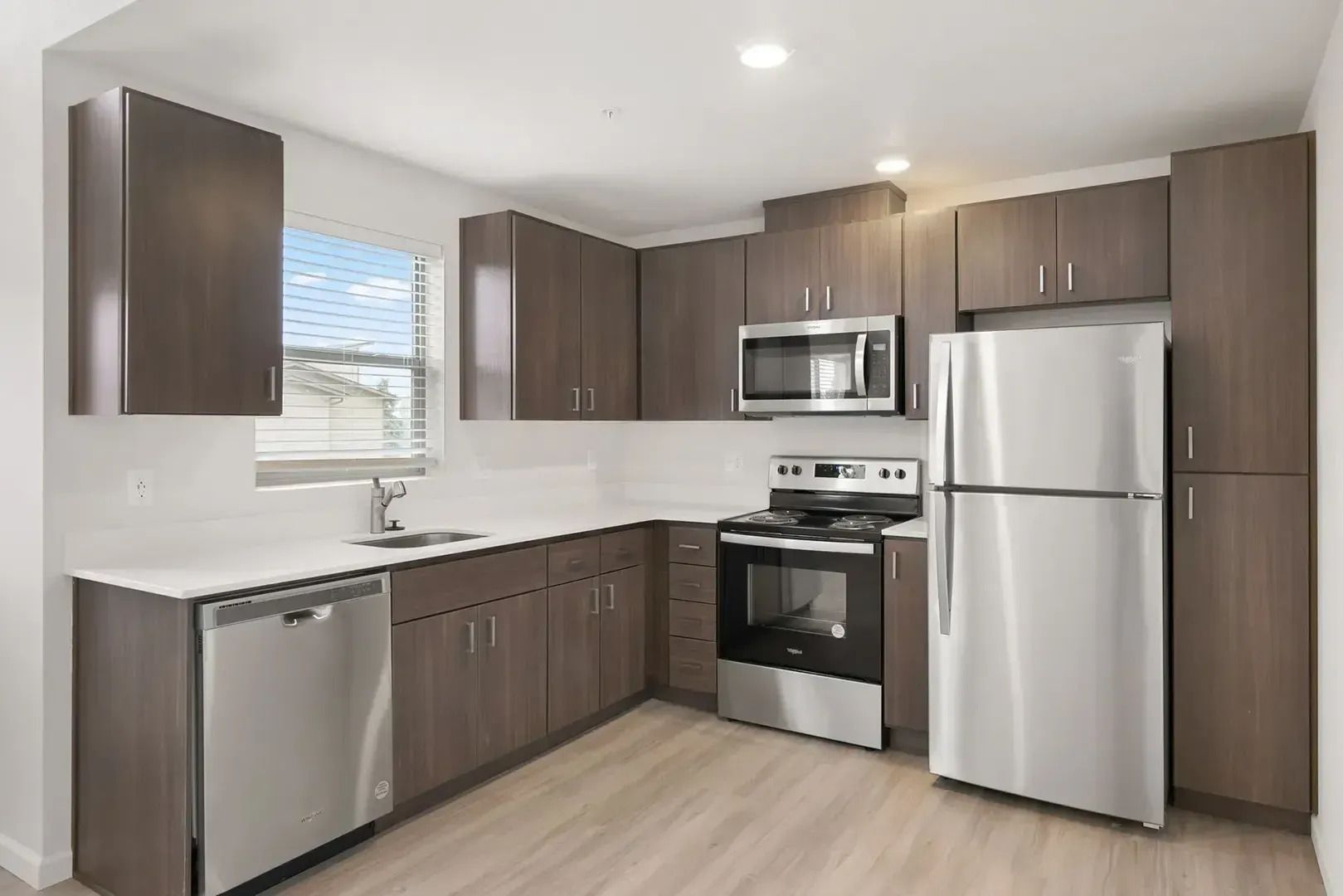Modern kitchen with stainless steel appliances, dark wood cabinets, white countertops, and a window over the sink