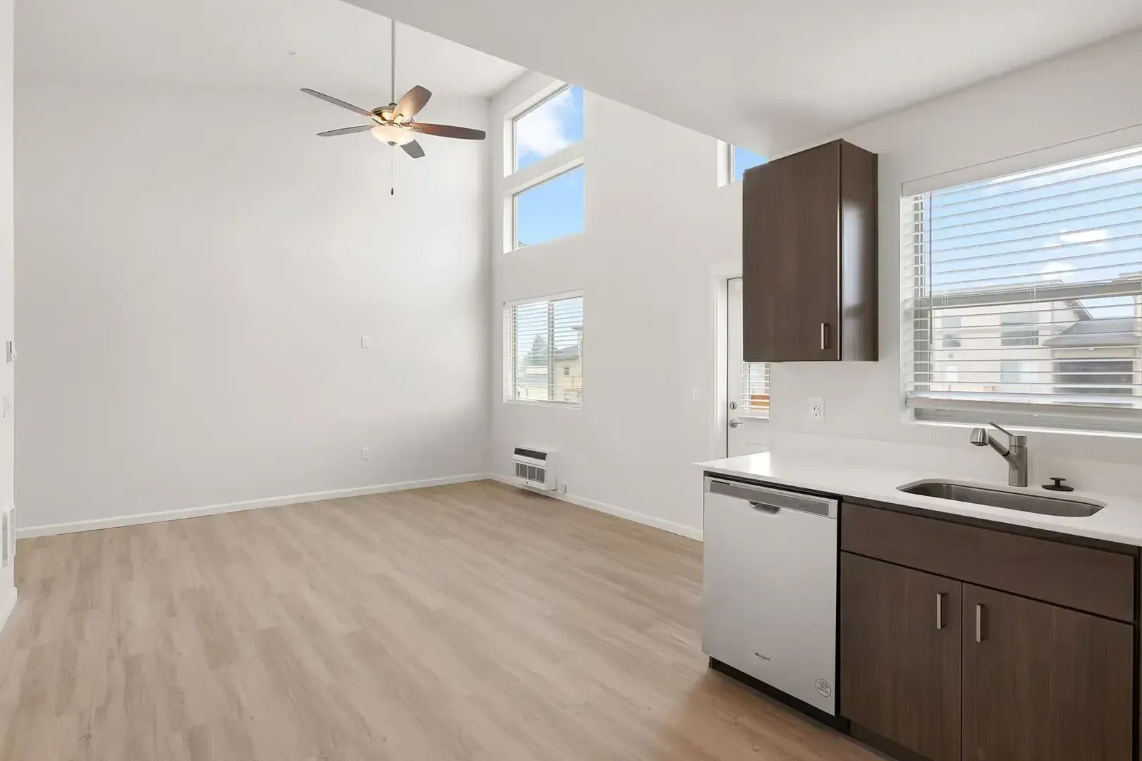 Empty modern kitchen and living area with light wood floors, white walls, and large windows.