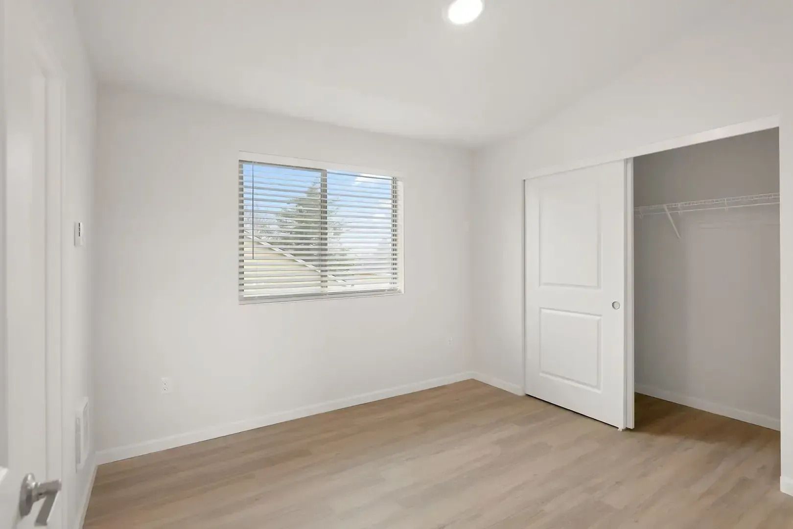 Empty white bedroom with wood flooring, window blinds, and an open closet door