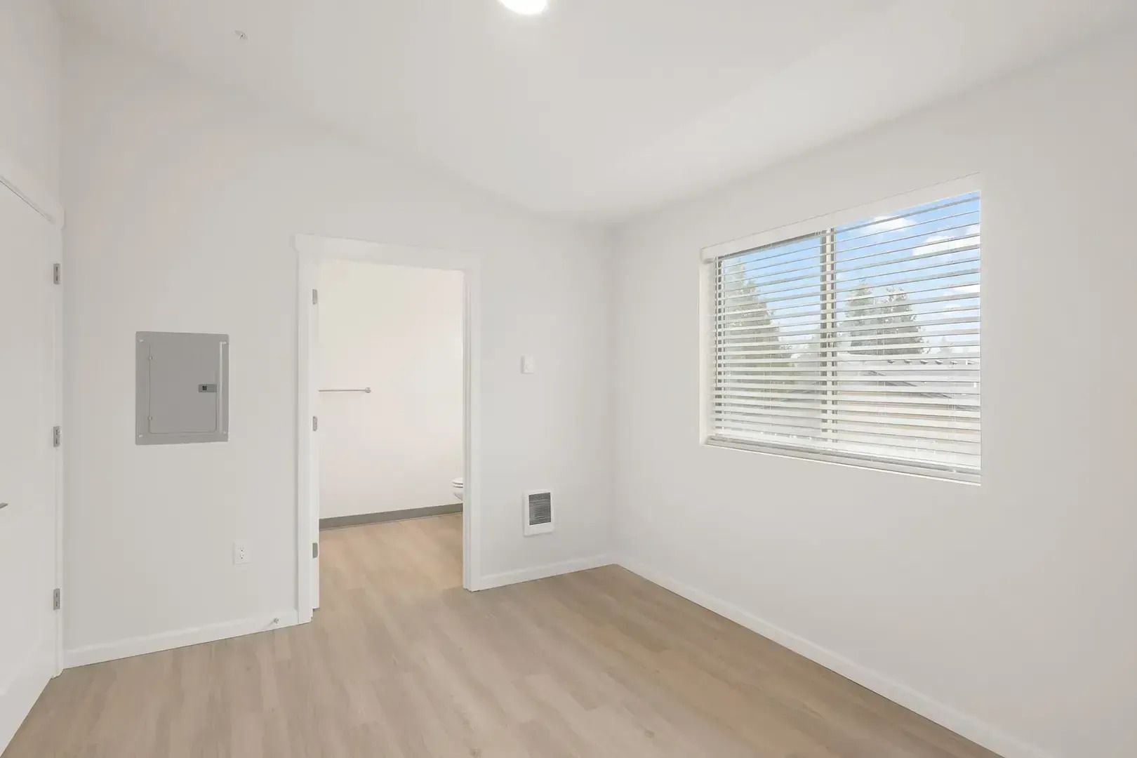Empty white room with light wood floor, window blinds, and an open doorway into another room