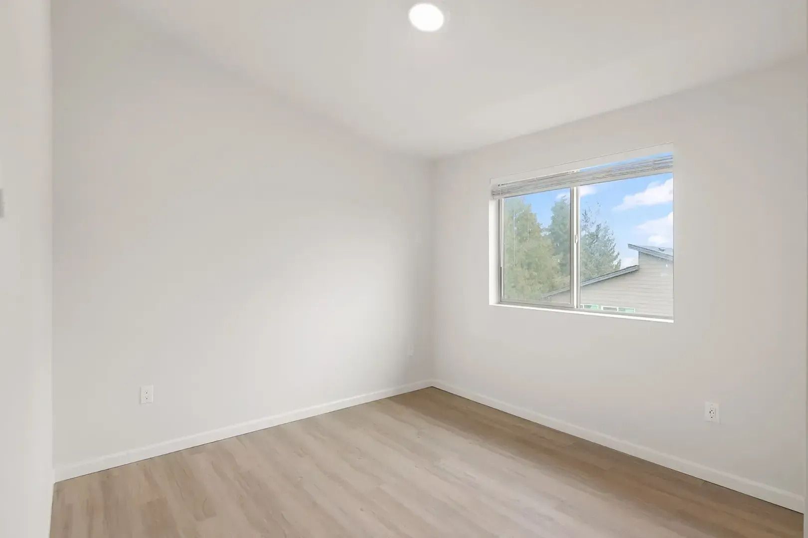 Empty white room with light wood floors and a window showing trees outside