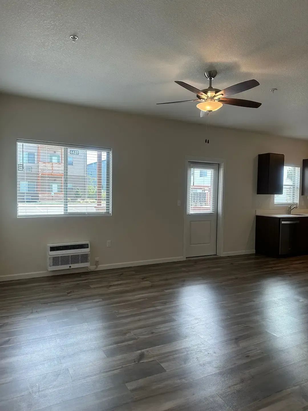 Empty living room with hardwood floors, ceiling fan, window, and door beside a small kitchen area