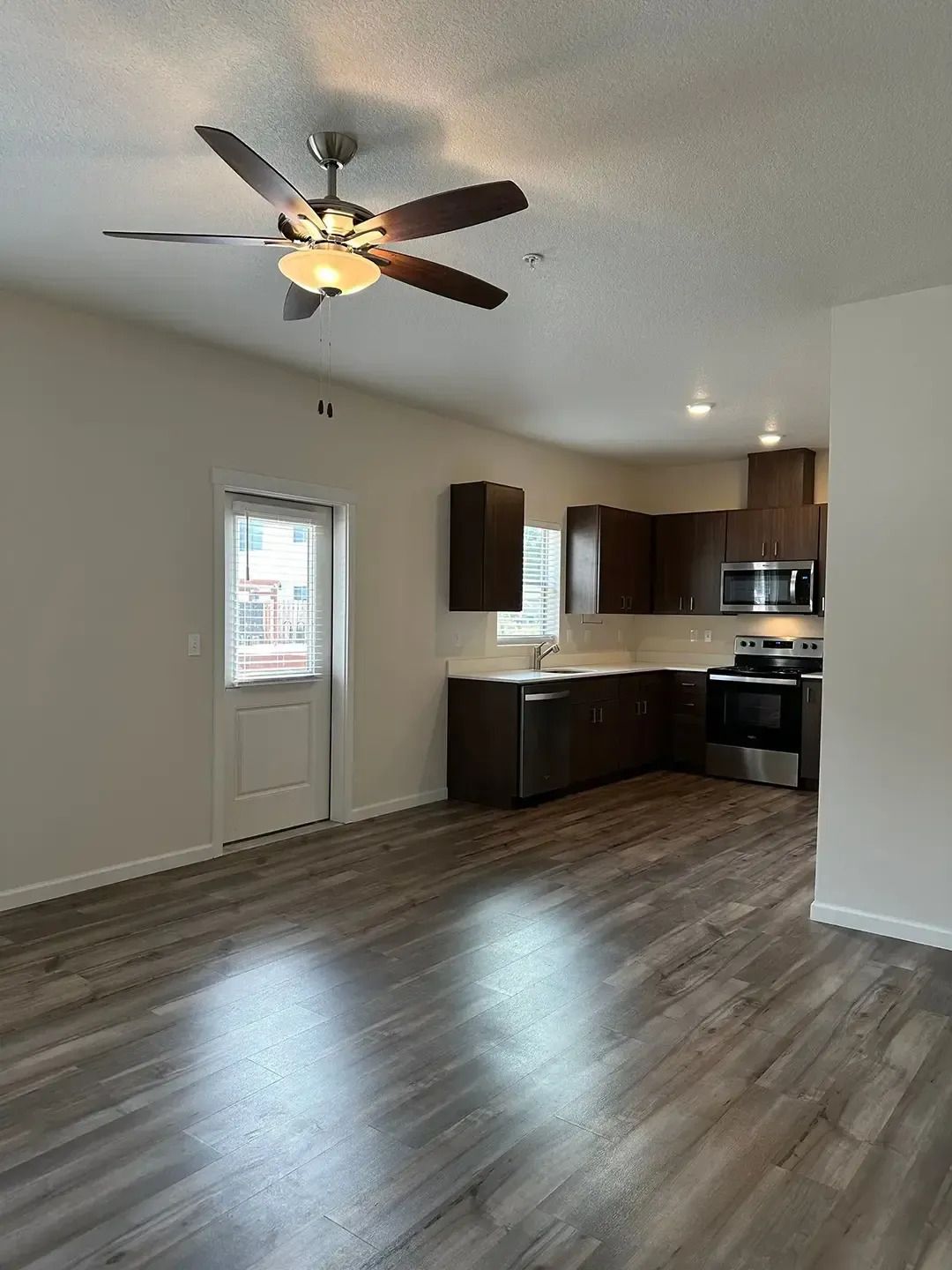 Empty open-plan room with gray wood floors, ceiling fan, and compact kitchen area.