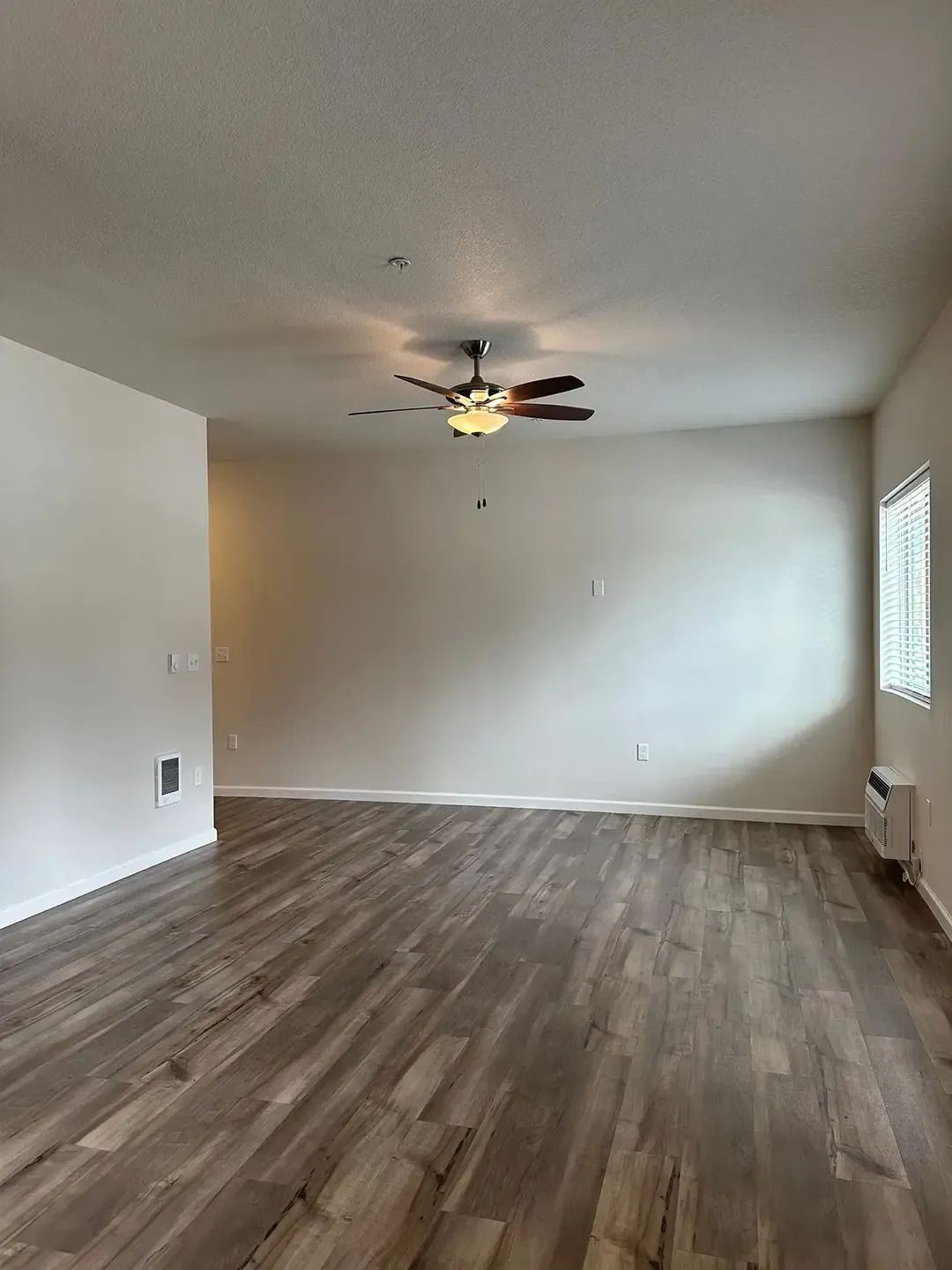 Empty living room with wood-look flooring, white walls, ceiling fan, and a small window on the right