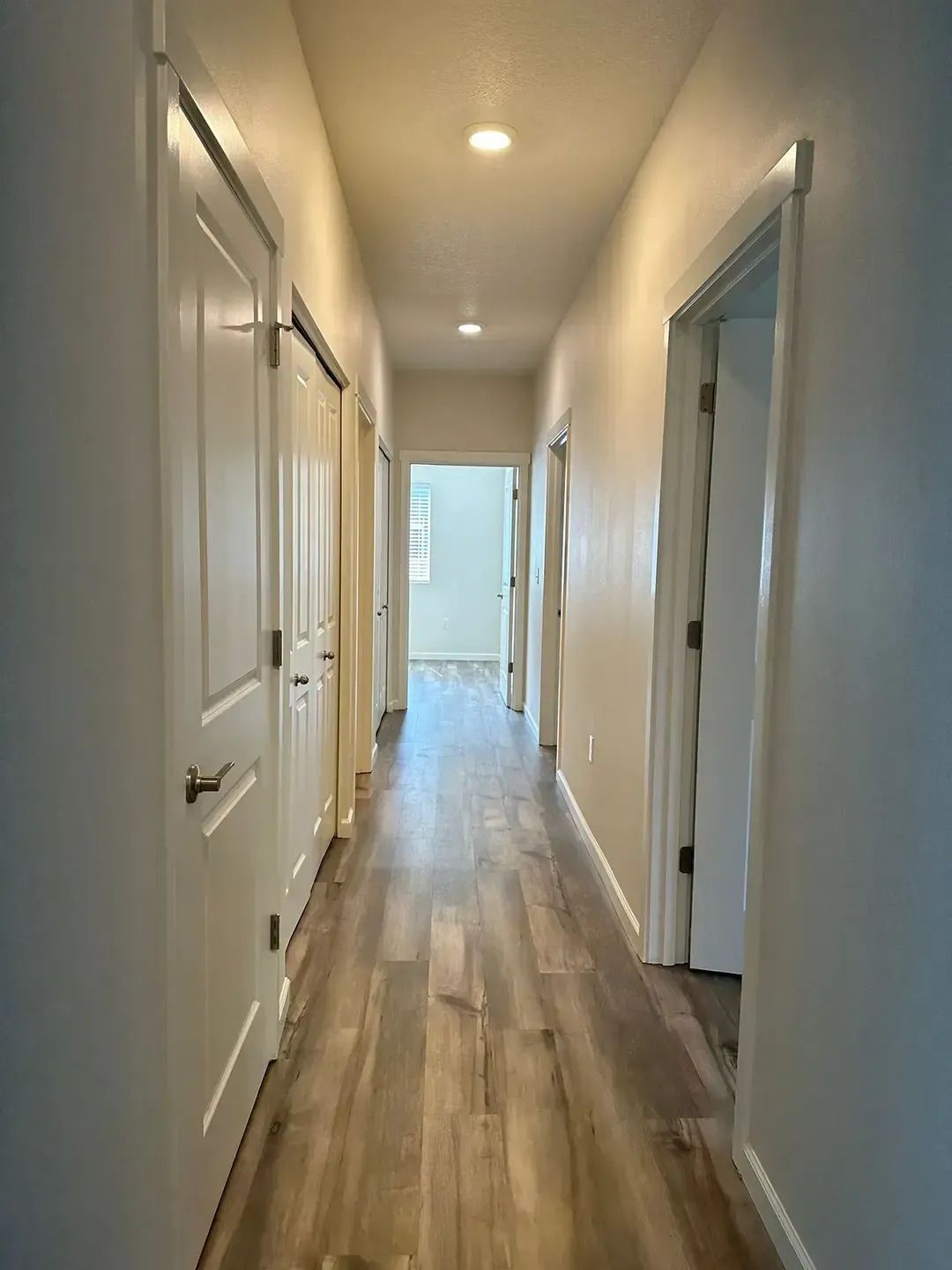 Narrow hallway with white doors, wood floors, and recessed ceiling lights leading to a bright room.