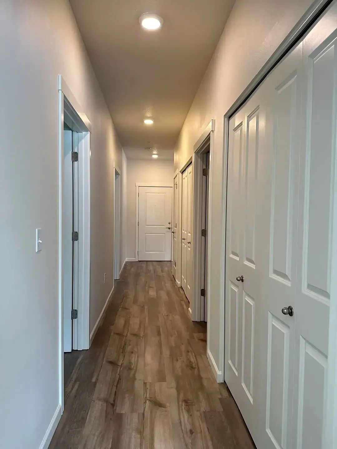 Narrow white hallway with wood flooring, ceiling lights, and multiple doors on both sides