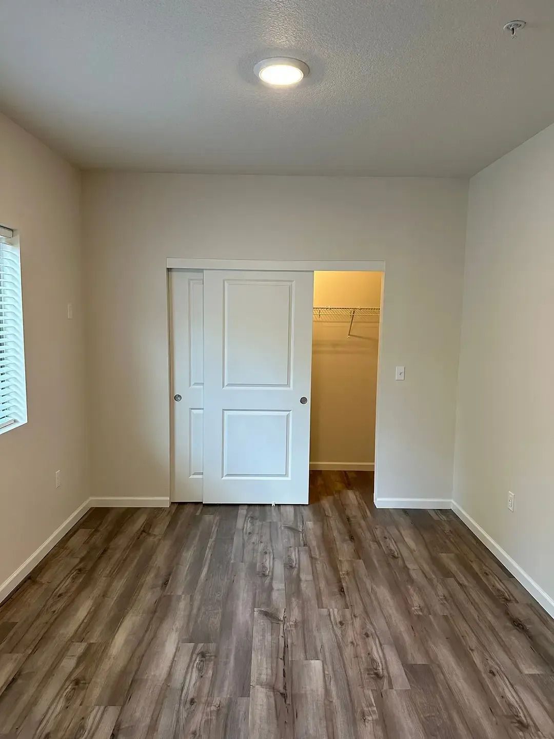 Empty beige room with wood flooring, white closet doors, and an open doorway to a lit hall.