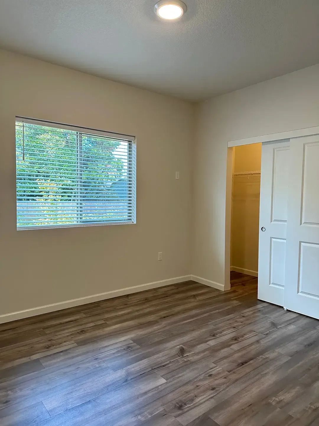 Empty beige room with wood floor, window blinds, ceiling light, and open closet door