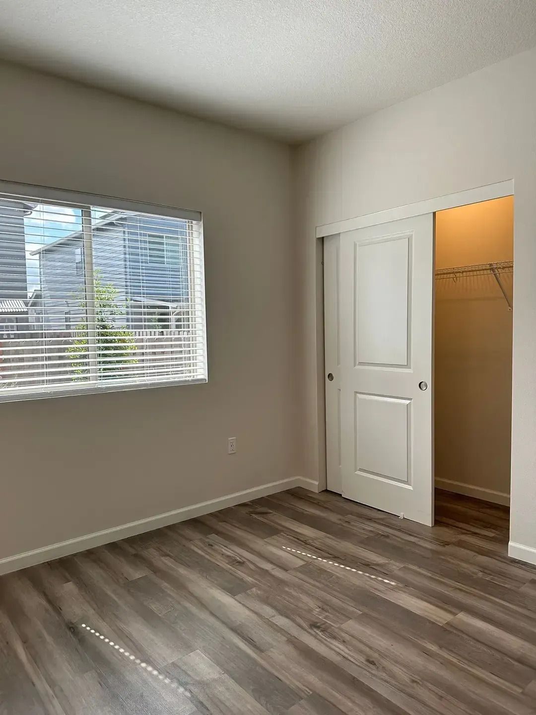 Empty bedroom with gray wood flooring, white walls, a window, and an open closet door.
