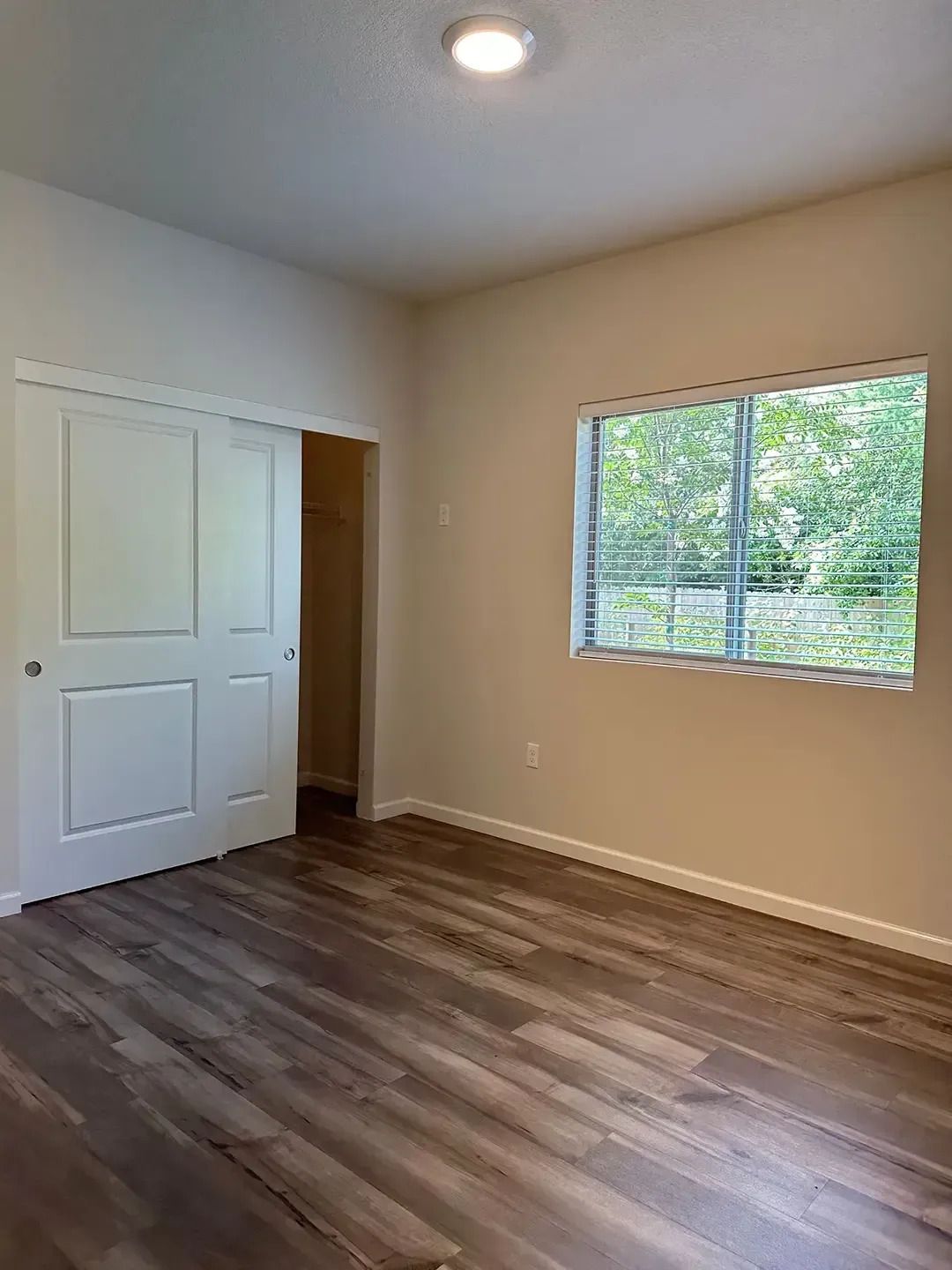 Empty beige bedroom with wood-look floors, closet doors, and a window.