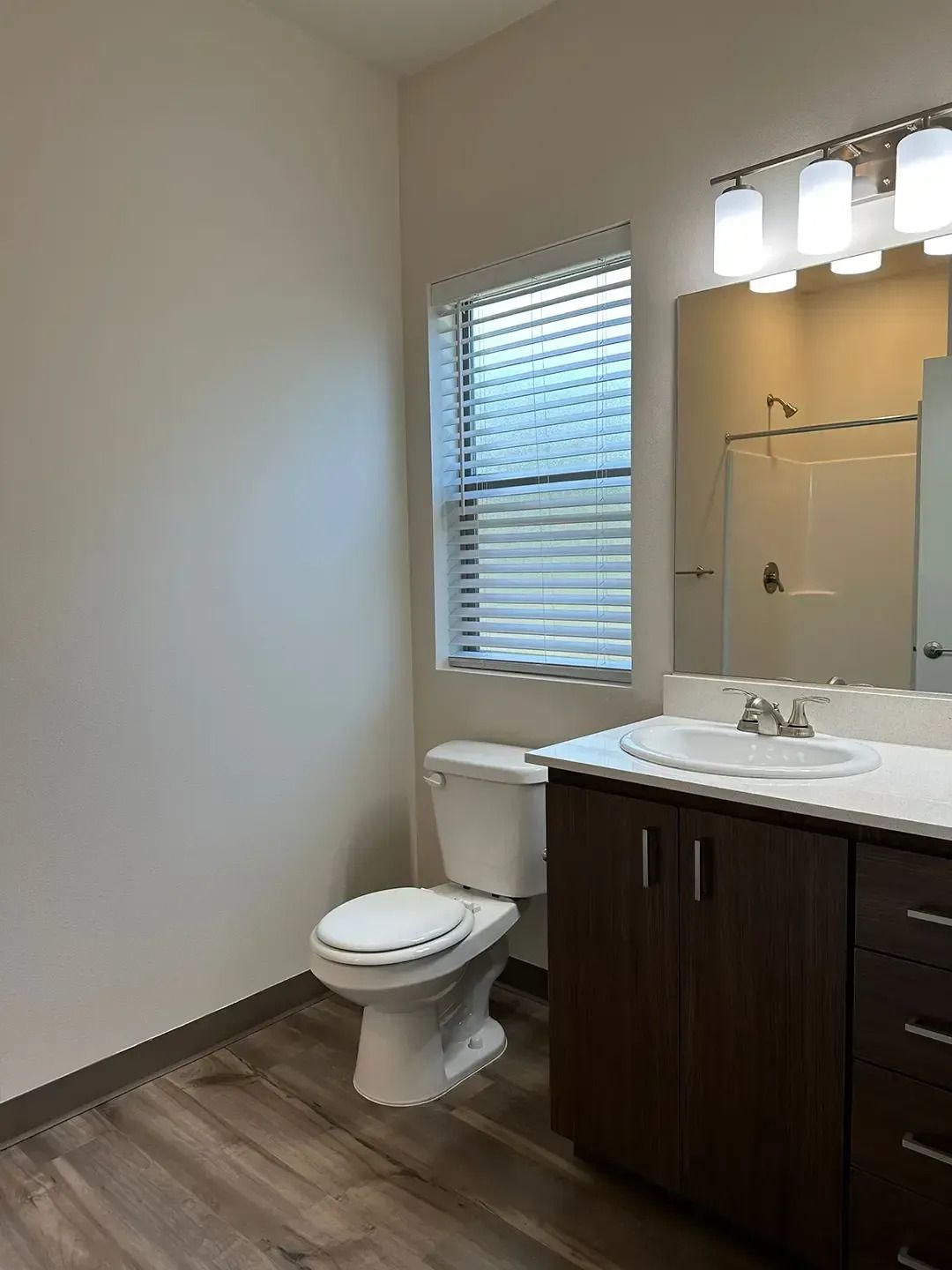 Bathroom with white toilet, dark vanity, mirror, and window blinds on a beige wall.