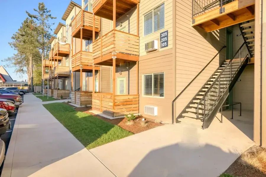 Apartment building exterior with balconies, stairs, and a sidewalk beside parked cars