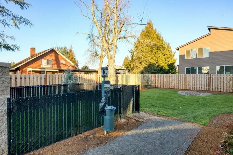Backyard with grass, fence, trees, and a paved path beside a black railing and blue post