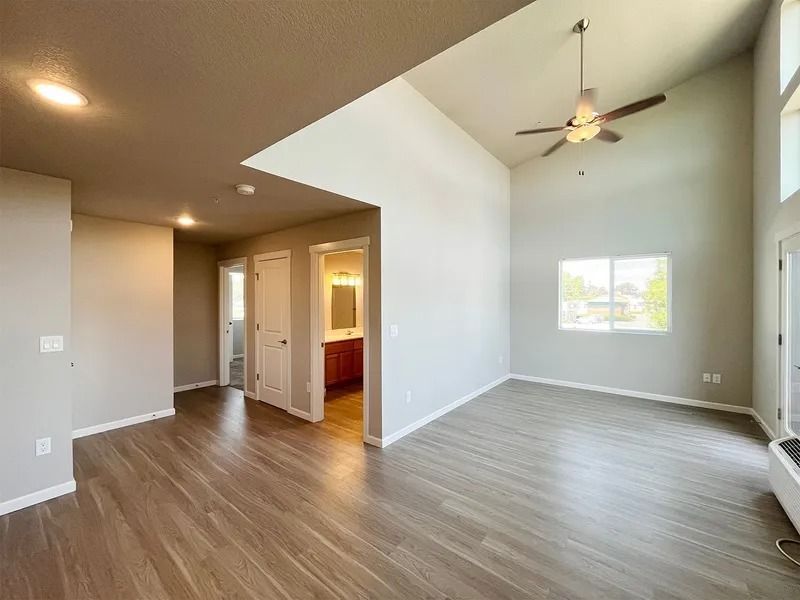 Empty modern living room with wood flooring, vaulted ceiling, ceiling fan, and a view into the kitchen.