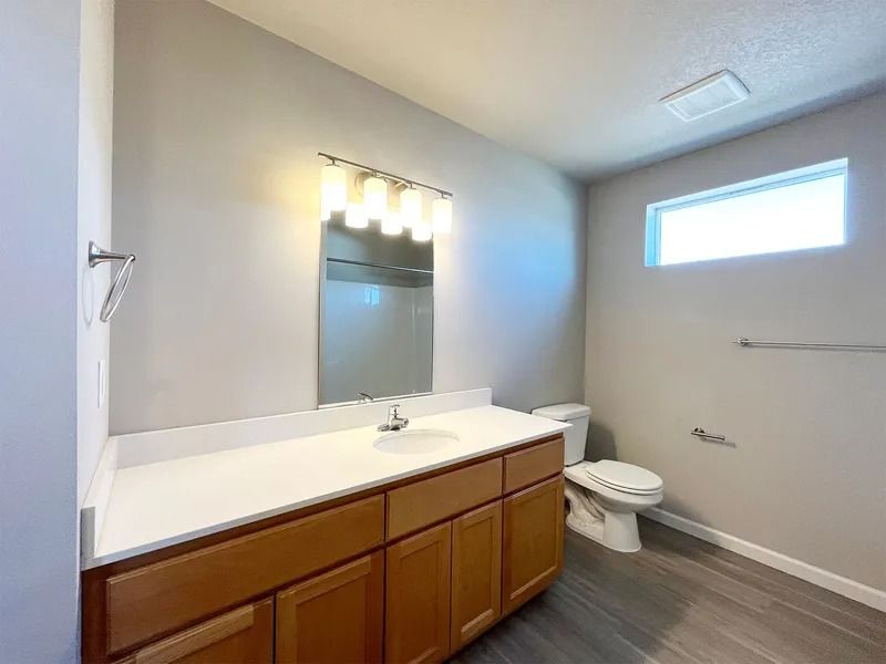 Bathroom with wood vanity, white countertop, mirror lights, toilet, and small high window.