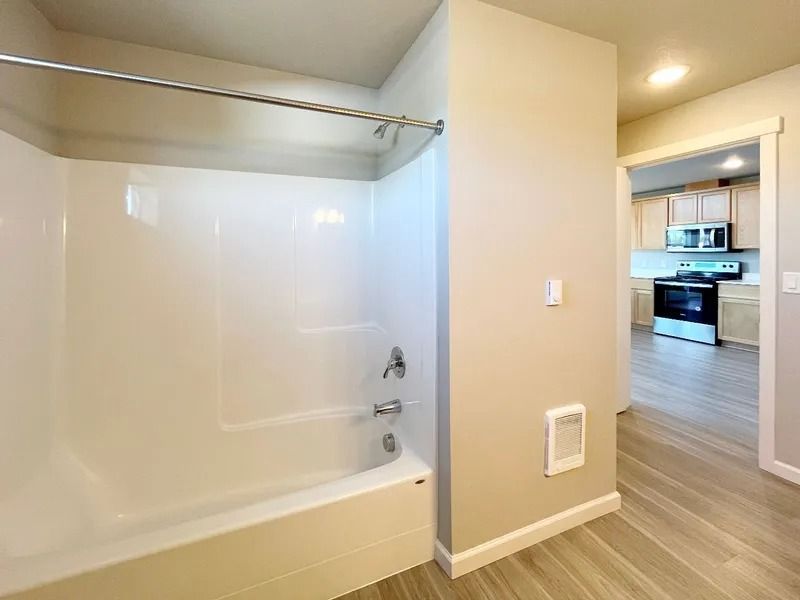 White bathroom with bathtub and shower, opening to a hallway and kitchen beyond.