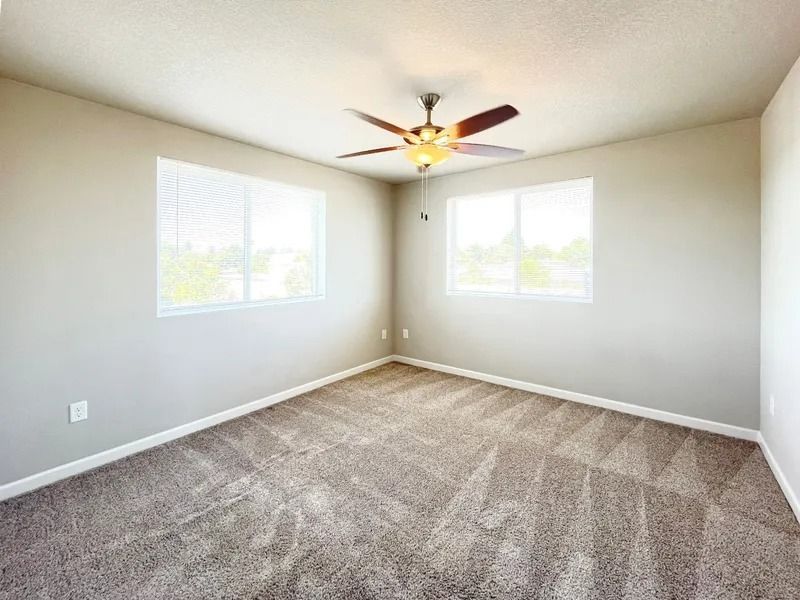 Empty carpeted bedroom with two windows and a ceiling fan, painted light beige.