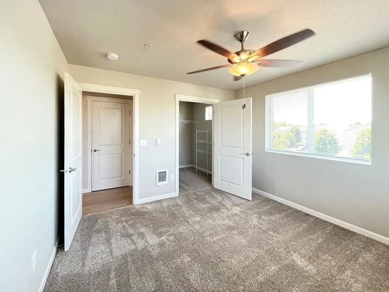 Empty bedroom with beige carpet, ceiling fan, open closet, and window letting in daylight
