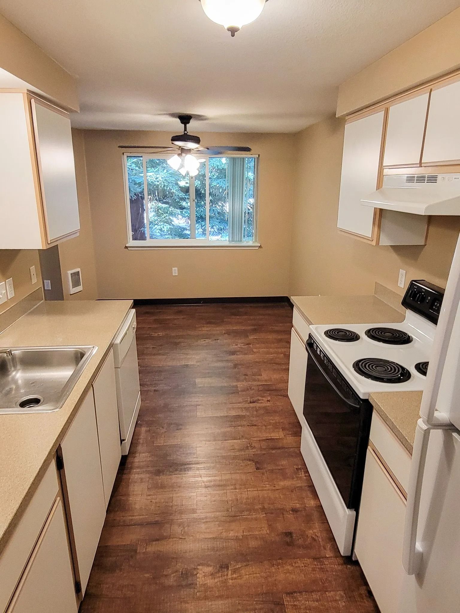 Narrow galley kitchen with white cabinets, brown floor, black stove, sink, and window at the end.