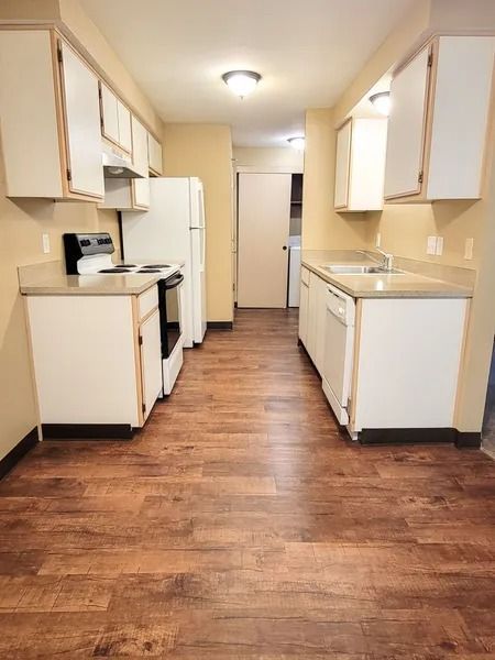 Empty narrow kitchen with white cabinets, wood-look floor, and refrigerator at the end.