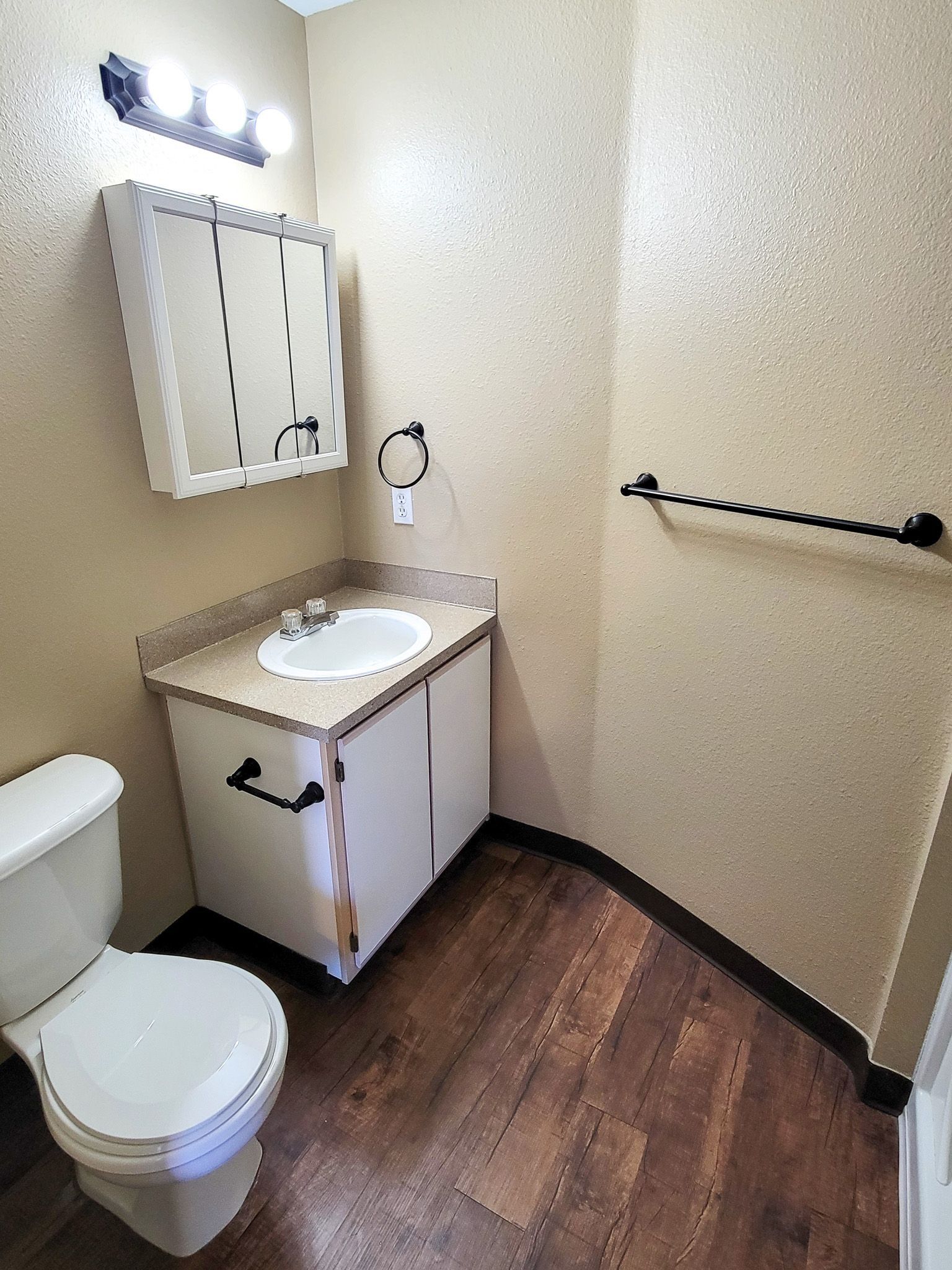 Small beige bathroom with toilet, vanity sink, mirror, and towel bar over dark wood floor.