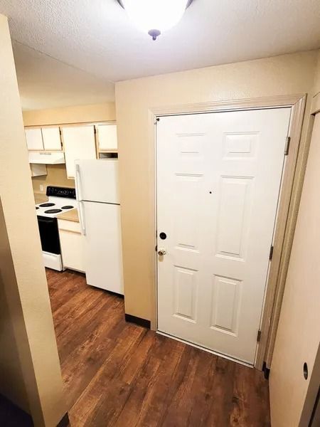 Small apartment entryway with white door, wood floor, and kitchen visible to the left.