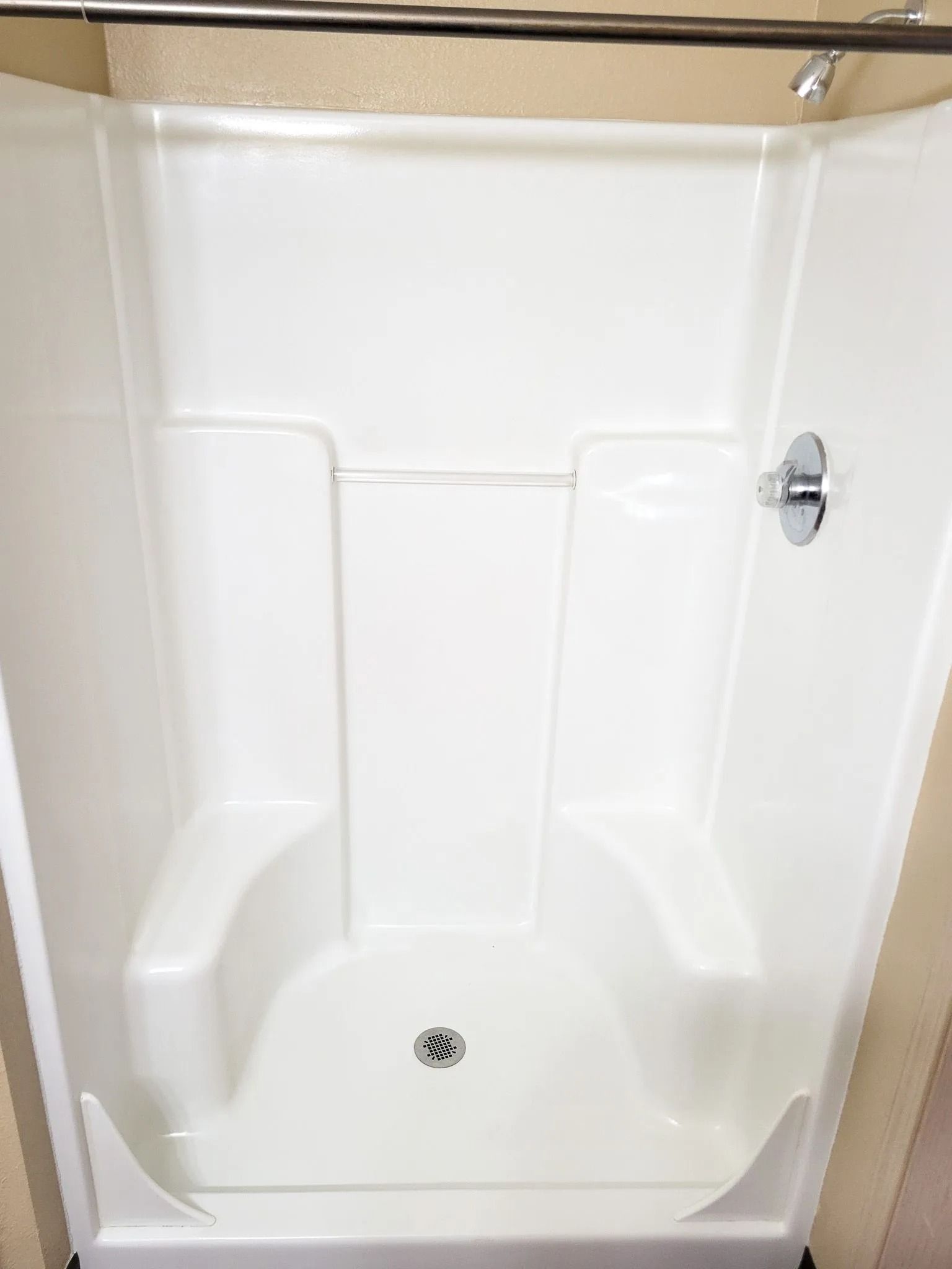 White shower stall with a built-in seat, grab bar, and drain on a beige wall background